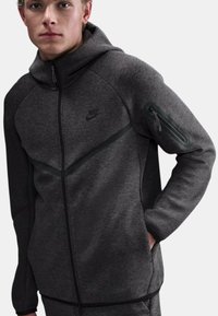 Giacca con cappuccio Nike in grigio scuro, con zip frontale, tessuto strutturato, tasca con zip sulla spalla e dettagli neri nel design.