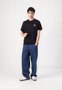 Black cotton t-shirt with logo, paired with loose-fitting blue denim jeans. Accessories include a black strap bag and white sneakers.