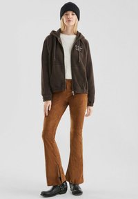 Bruine fleece zip-up hoodie met voorzakken, witte letters, gecombineerd met bruine flared broeken met zijsplitten en zwarte enkelboots.