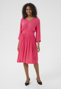 Robe rose avec un décolleté en V et des manches trois-quarts. Présente une taille smockée et une jupe évasée avec une texture douce et fluide. Talons aiguilles noirs.