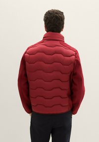 Czerwona pikowana kurtka z wysokim kołnierzem i strukturalnym wzorem fal, z podgumowanymi bocznymi panelami i gładką fakturą.