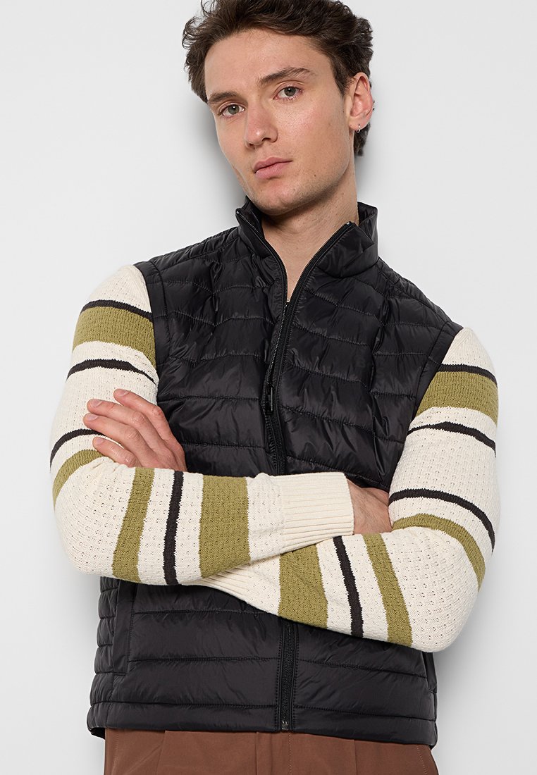 Gilet nero invernale con colletto alto, chiusura con zip e design trapuntato orizzontale, indossato sopra un maglione crema con righe verdi.