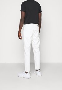 Białe dżinsy o prostym kroju, pięć kieszeni, z czarną metką w pasie, noszone z czarnym t-shirtem z podwiniętymi rękawami i białymi trampkami.