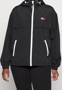 Svart lättviktsjacka i slät syntetisk material, med dragkedja framtill, elastiska ärmslut och en logopatch på bröstet.