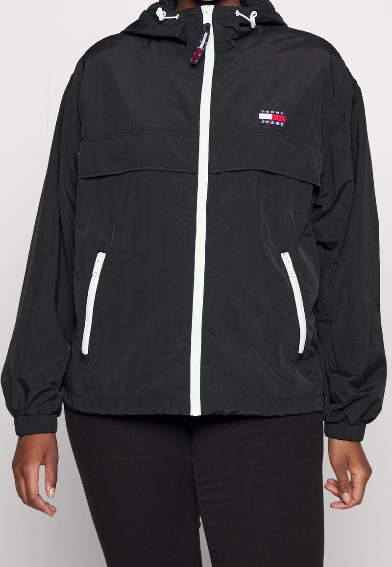 Svart lättviktsjacka i slät syntetisk material, med dragkedja framtill, elastiska ärmslut och en logopatch på bröstet.