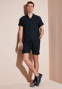 Cămașă scurtă cu mânecă, de culoare albastru închis, cu un model texturat, asociată cu pantaloni scurți asortați de aceeași nuanță și pantofi sport cu accente gri, pe un fundal neutru.