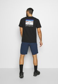 Camiseta de algodón negra con un gráfico de montaña en la parte posterior, combinada con pantalones cortos azules y calcetines negros, vista desde atrás.