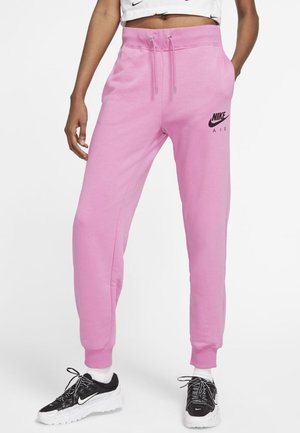 Personne portant un pantalon de survêtement rose Nike Air avec une taille à cordon, des chaussettes blanches et des baskets Nike noires sur un fond blanc uni.