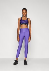 Lila sportmelltartó fekete logós pánttal, amelyet lila magas derekú leggingssel kombináltak. A modellen fekete sportcipő van. Sima textúra.