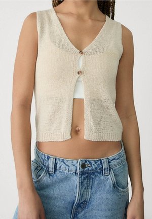 Beżowa dzianinowa kamizelka bez rękawów z dwoma guzikami, noszona na białym crop topie, w zestawie z jasnoniebieskimi dżinsami o wysokim stanie.