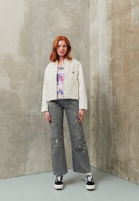 Vit kort jacka, blommig grafik t-shirt, grå vida byxor med färgstänk och svarta sneakers med vita sulor. Slät textur.