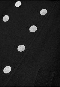 Tissu noir avec deux rangées verticales de boutons argentés brillants présentant des profils en relief, et une poche cousue visible en bas à droite.