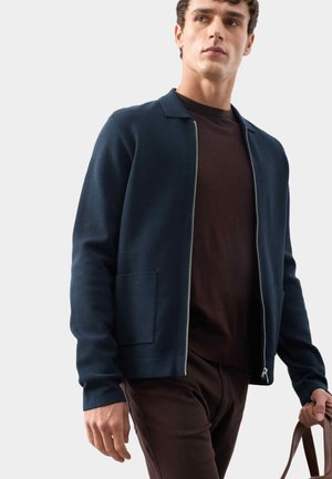 Mand iført en mørkeblå zip-up jakke over en brun skjorte og bukser, holder et brun lædertaske håndtag mod en lys grå baggrund.