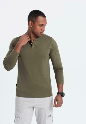 Maglietta henley a maniche lunghe verde oliva con placca a bottoni nera, tessuto strutturato e design aderente. Abbinata a pantaloni cargo chiari.