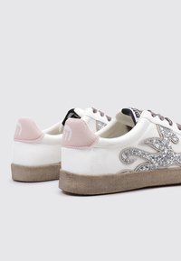 Baskets blanches avec un talon texturé rose clair, accent en paillettes argentées et semelle en caoutchouc beige. Elles présentent des lacets et un branding discret sur le talon.