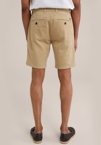 Pantaloni scurți din bumbac bej, cu croială dreaptă, două buzunare la spate cu butoane închise la culoare și margini răsucite. Purtați cu încălțări slip-on din material textil închise la culoare.