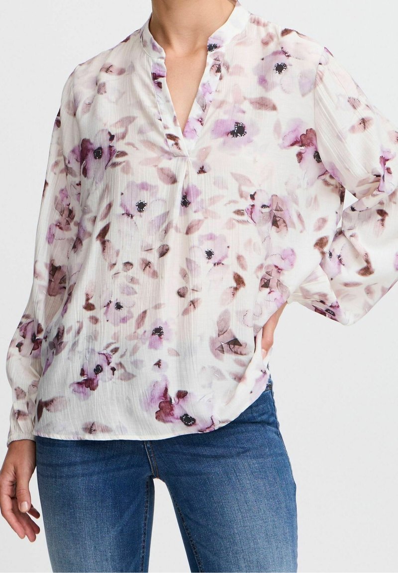 Blouse fluide en tissu léger et texturé, affichant une base blanche avec un motif floral rose et violet, et un col mandarin.
