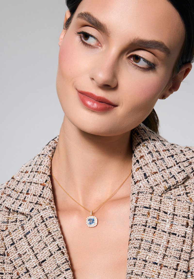 Złoty naszyjnik z wisiorkiem w okrągłym kształcie, z centralnym niebieskim kamieniem otoczonym drobnymi białymi kamieniami, noszony na teksturowanym, jasnym żakiecie.
