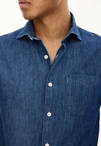 Donkerblauwe denimhemd met een knoopsluiting, kraag en een enkele opgestikte zak. Heeft een zachte textuur en subtiele variatie in de stof.