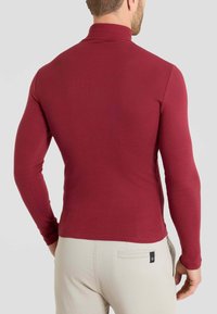 Maglietta a maniche lunghe color marsala con colletto alto, texture liscia, design aderente e cuciture a contrasto; indossata con pantaloni di colore chiaro.
