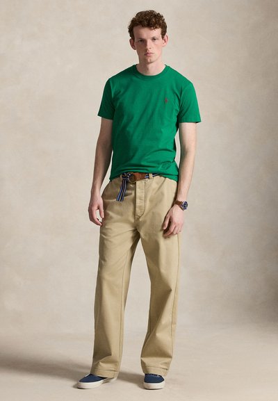 Ung mand står iført grøn kortærmet skjorte, beige bukser med vide ben og en stribet bælte, blå slip-on sko og et armbåndsur.