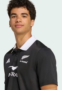Sort polo shirt med hvid krave, præget mønster og logoer for "All Blacks" og "Adidas" på forsiden. Korte ærmer.