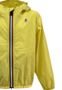 Giacca impermeabile gialla con cappuccio, zip frontale completa e polsini elasticizzati. Presenta un logo sul petto e dettagli a strisce a contrasto sulla zip.