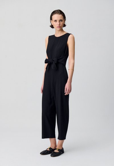 Combinaison noire avec un corps ajusté, sans manches, et un nœud à l'avant à la taille. Pantalons courts amples et texture douce.