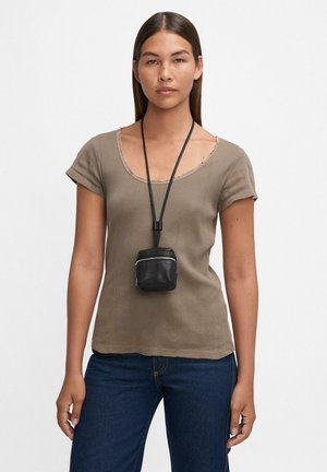 Femme portant un haut taupe à manches courtes et un jean bleu foncé avec une petite pochette en cuir noir suspendue à une longue bandoulière autour du cou.