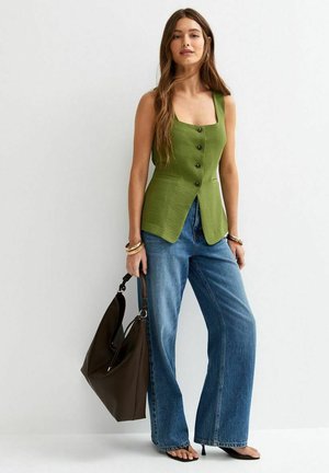 Femme portant un haut vert sans manches à boutons et un jean bleu à jambes larges, tenant un grand sac à main marron, debout contre un mur blanc.