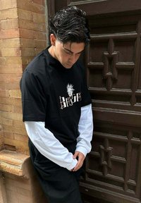 Camiseta gráfica negra superpuesta sobre una camisa de manga larga blanca, con un diseño audaz. La lleva puesta con pantalones negros, de pie contra una pared texturizada.