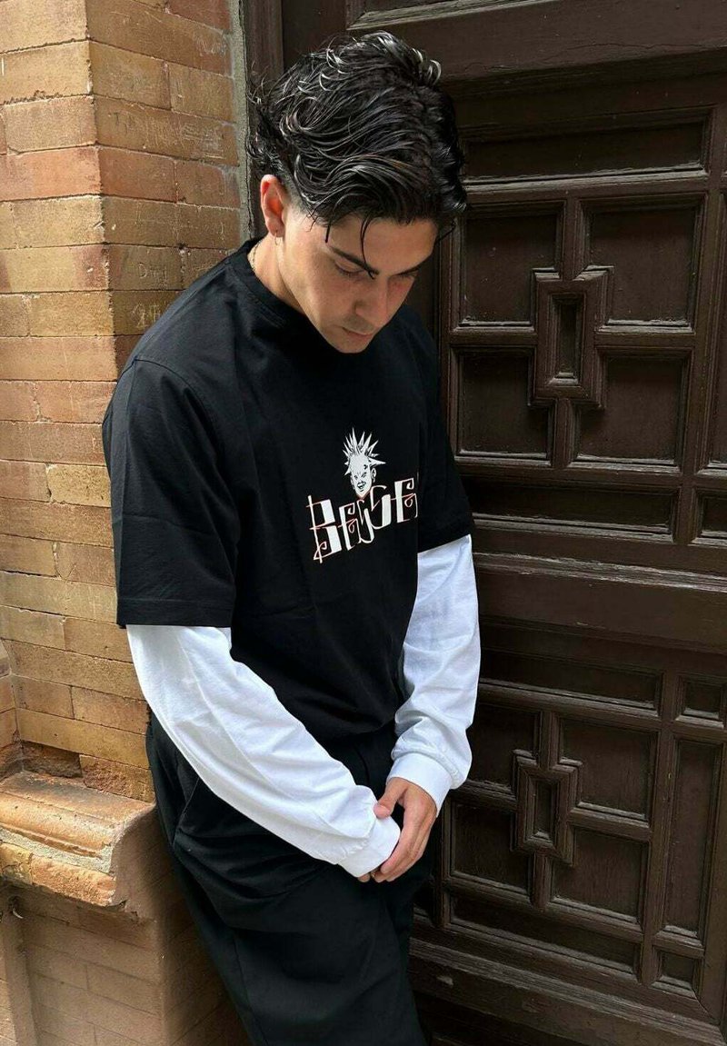 Camiseta gráfica negra superpuesta sobre una camisa de manga larga blanca, con un diseño audaz. La lleva puesta con pantalones negros, de pie contra una pared texturizada.