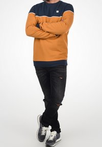Sweat-shirt bicolore orange et marine avec un col rond, des manches longues et des poignets côtelés, assorti à un jean noir déchiré et des baskets grises.