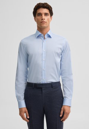 Mann, der ein hellblaues langärmeliges Hemd trägt, das in eine dunkelblaue maßgeschneiderte Hose gesteckt ist, vor einem schlichten Hintergrund.