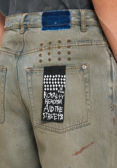 Heledad denims kerge rohelise värviga, sinise värvitooni plaastriga, kuldvärvi ristikangaga tikandiga ning musta kangast sildiga, millel on valge tekst graffiti stiilis.