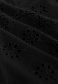 Tissu noir avec broderie florale ajourée disposée en rangées diagonales, montrant la texture et de petits détails découpés.