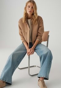 Beige jacket with two front pockets, striped sweater underneath, wide light blue jeans, and beige sneakers, seated on a metal chair.