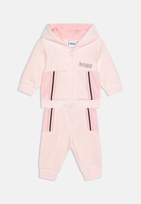 Ensemble en velours rose comprenant un hoodie zippé avec un logo, des accents rayés, et un pantalon coordonné avec des poches latérales.