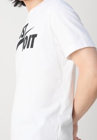 Vit bomulls t-shirt med korta ärmar, med en svart "JUST DO IT" logotyp på framsidan. Slät textur, standard rund halsdesign.