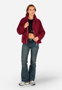 Burgundi fliisjakker, millel on krae, lukuga sulgemine ja soonikkoes, koos laienevate siniste teksade ja hõbedaste spordijalatsitega.