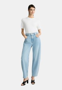 Jeans in denim leggero blu chiaro con vestibilità rilassata, vita alta, due tasche laterali e chiusura con bottone, abbinati a una t-shirt bianca aderente e tacchi neri.