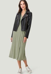 Zwarte leren motorjack met rits- en drukknoopdetails, gelaagd over een groene jurk met een geometrisch patroon en een midi-lengte.