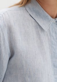 Ljusblå skjorta med krage och ett fint vertikalt randmönster. Tyget verkar ha en textur, med en knappstängning nära kragen.