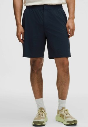 Homme portant un short de sport bleu marine foncé, associé à des chaussettes blanches montantes et des chaussures de course beiges, debout sur un fond uni.