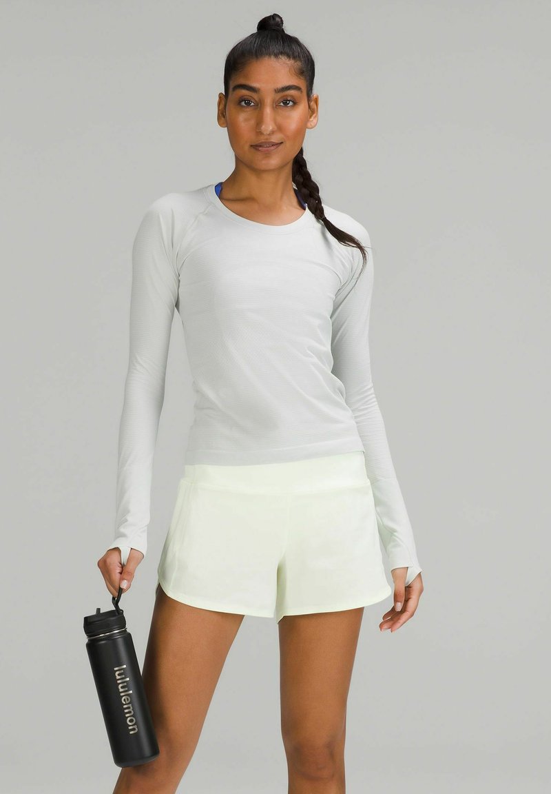 Haut de sport à manches longues gris clair en tissu texturé, complété par un short jaune néon et une bouteille d'eau noire avec un logo blanc.