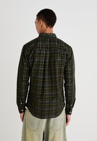 Green plaid shirt with a buttoned collar and long sleeves, featuring a soft texture and a curved hem, worn with light-colored trousers.