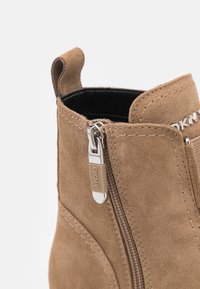 Bottine en daim beige avec fermeture éclair latérale, munie d'une tirette argentée gravée "DKNY" et d'un panneau élastique marqué sur la partie supérieure.