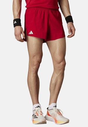 Athlète masculin portant un short rouge, des bracelets noirs et des chaussures de course blanches avec des touches orange, debout les jambes légèrement écartées.