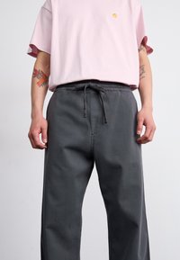 T-shirt en coton rose clair avec un petit logo jaune, associé à un pantalon à cordon gris foncé avec des poches latérales et une coupe décontractée.