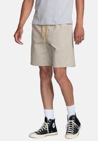 Shorts légers beiges avec une taille élastique et un cordon orange. Portés avec des baskets montantes noires et des chaussettes blanches.